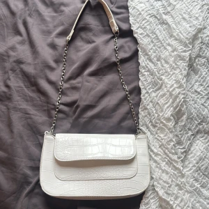 Vit axelväska med kedja och croco-mönster - Snygg vit axelväska med croco-mönstrad yta och silverfärgad kedja som axelrem. Väskan har ett stilrent lock med magnetknäppning och ett rymligt fack inuti. Perfekt storlek för det viktigaste och ger en trendig touch till din outfit.