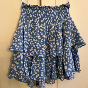 Blommig blå volangkjol från Samsa - Supersöt blå kortkjol från Samsa Collection med vitt blommigt mönster. Kjolen har flera volanger och smock i midjan för en bekväm passform. Perfekt för dig som gillar en feminin och lekfull stil. Materialet känns lätt och luftigt. Köpt på Ullared 