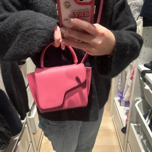 Rosa handväska från ATP Atelier - Snygg rosa handväska från ATP Atelier i skinn med unik asymmetrisk lockdesign. Väskan har både handtag och axelrem i samma färg, samt guldfärgade metalldetaljer. Perfekt storlek för det viktigaste och ger en poppig touch till din outfit.