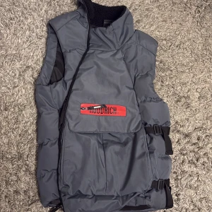 Grå dunväst från hoodrich - Snygg grå dunväst från Woolrich med stor ficka framtill och röd logotypdetalj. Västen har justerbara spännen i sidan och hög krage med svart fleecefoder. Perfekt för lager-på-lager och streetstyle.