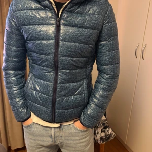 Blå pufferjacka från Terranova L - Säljer en mörkblå pufferjacka från Terranova i storlek L. Jackan har huva, horisontella sömmar och dragkedja framtill. Materialet är syntet och jackan är vadderad för extra värme. Perfekt för kyliga dagar och har en clean, sportig look.