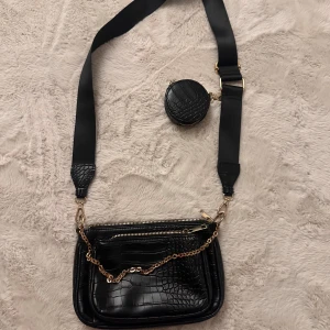 Svart axelväska med kedja och croco mönster - Snygg svart axelväska med croco mönster och guldfärgad kedjedetalj. Väskan har dragkedja upptill och en bred axelrem med matchande liten rund väska. Köpt från New yorker!!