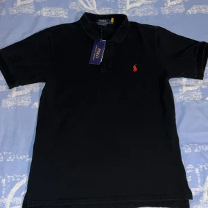 Svart pikétröja från Polo Ralph Lauren - Klassisk svart pikétröja från Polo Ralph Lauren med röd broderad logga på bröstet. Tröjan har korta ärmar, krage och knappar framtill. Tillverkad i bomull för en skön och stilren känsla. Perfekt för en clean och tidlös look.