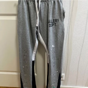 Grå mjukisbyxor Gallery Dept. - Grå mjukisbyxor från Gallery Dept. med svart logga på vänster lår, vita och svarta detaljer längs benen samt färgstänk som ger en cool streetkänsla. Byxorna har snörning i midjan och är tillverkade i mjukt bomullsmaterial.