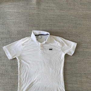 Vit golfpikétröja från Helly Hansen - Vit pikétröja från Helly Hansen med klassisk krage och knappar framtill. Tröjan har korta ärmar och HH-logga broderad på bröstet. Perfekt för en clean och sportig look, tillverkad i mjuk bomull som är skön mot huden.