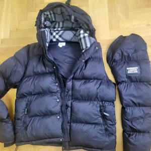 Svart dunjacka från Burberry - Säljer en svart pufferjacka från Burberry London England med avtagbara ärmar. Jackan har dragkedja, knappar och klassiskt rutigt foder i huvan. Perfekt för kalla dagar och riktigt snygg streetstil.
