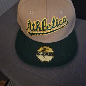 Oakland Athletics keps från New Era - Snygg 59FIFTY keps från New Era med Oakland Athletics broderat i gult och grönt framtill. Kepsen är beige med mörkgrön skärm och har officiella MLB-loggor på baksidan och insidan. Tillverkad i 100% polyester, perfekt för dig som gillar baseball och streetwear.