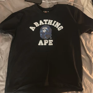 Svart BAPE t-shirt med blå camo - Svart t-shirt från A Bathing Ape med klassiskt tryck på bröstet. Stor vit text och ikoniskt BAPE-huvud i blått camo. Rund hals och korta ärmar. Perfekt för dig som gillar streetwear och vill sticka ut med en ikonisk design.