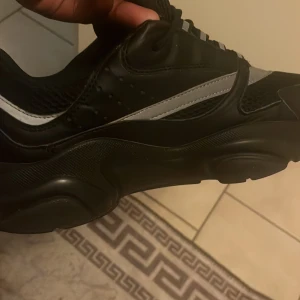 Svarta Dior sneakers med vita detaljer - Säljer ett par chunky svarta Dior sneakers i storlek 42 med vita detaljer och meshpartier. Skorna har en kraftig sula, snörning och coola kontraster mellan skinn och mesh. Perfekta för dig som vill sticka ut med en lyxig streetstyle.