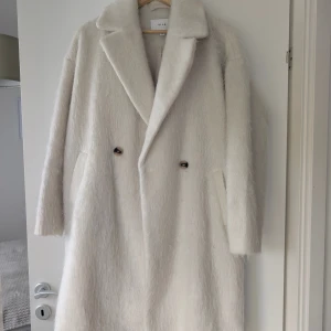 Vit fluffig kappa från Vila - Säljer en lång, vit kappa från Vila med fluffig och mjuk känsla. Kappan har bred krage, dubbelknäppning med två knappar framtill och diskreta fickor på sidorna. Perfekt för dig som vill ha en stilren och mysig look under kyliga dagar.