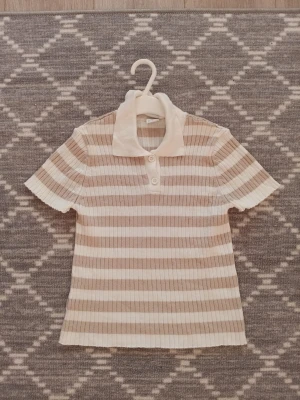Randig ribbad topp från Lindex Kids - Säljer en kortärmad topp från Lindex Kids i storlek 134/140. Toppen är ribbad och har ränder i beige och vitt, med klassisk krage och två knappar framtill.🧸❤️ Materialet är mjukt och stretchigt, perfekt för en chill look. Denna var min favorit länge! 50 kr+frakt💰