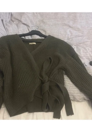 Olivgrön omlottkofta från Moocci - Säljer en olivgrön stickad omlottkofta från Moocci. Koftan har långa ärmar, bred ribbad struktur och knyts i midjan med ett brett band. Perfekt för lager på lager och ger en chill men ändå snygg look.