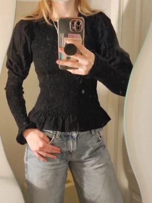 Svart blus med puffärm och smock  - Använd fåtal gånger! Svart blus med puffärm på övre delen av armen och smockad struktur över hela kroppen och ärmsluten. Blusen har en rund halsringning och volangkant nedtill. 