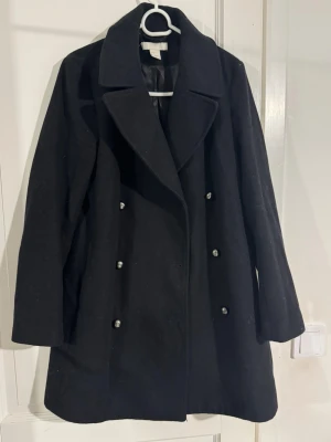 Svart rock med dubbelknäppning - Snygg svart rock från H&M  med bred krage och dubbelknäppning framtill. Jackan har en klassisk, rak passform och är gjord i ullblandning som håller dig varm under kyliga dagar. Perfekt för dig som gillar stilrena och tidlösa plagg.storlek 34