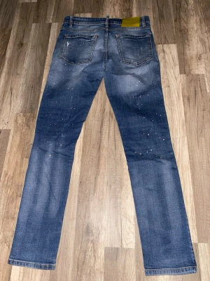 Dsquared2 blå jeans med slitningar - Säljer ett par blå Dsquared2 jeans med coola slitningar och patchade detaljer framtill. Jeansen har färgstänk och en klassisk femficksmodell med raka ben. Bak finns Dsquared2-logga på gult läderpatch. Perfekta för dig som gillar streetwear och vill sticka ut.