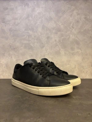 Axel Arigato Clean 90 - Snygga svarta Axel Arigato Clean 90 med vit platt sula och diskret logga på sidan och hälen. Skorna har rund tå, snörning och är tillverkade i slätt skinn med mockadetalj vid hälen. Perfekta för dig som gillar stilrena och minimalistiska sneakers. I bra skick men finns lite slitage från användning! Hälen har slitage se bild men inget som påverkar utseende eller funktion. 