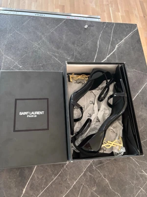 Svarta Saint Laurent sandaler med YSL-klack - Säljer ett par svarta sandaler från Saint Laurent med glansig finish och ikonisk guld YSL-klack. Skorna har smala remmar över tårna och runt ankeln, samt en fyrkantig tå. Perfekt för dig som vill sticka ut med lyxig design och detaljer.