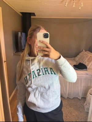 Hoodie från Axel Arigato  - Grå hoodie från Axel Arigato med grön broderad text framtill. Klassisk modell med huva, snörning och stor magficka. Mjuk bomullskvalitet och ribbade muddar vid ärmslut och nederkant. Perfekt för chill dagar och streetstyle.