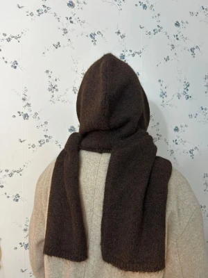 Mörkbrun stickad väst med huva - Unik mörkbrun stickad väst med stor huva och långa paneler som hänger ner över axlarna. Västen har en enkel, rak form och är tillverkad i ett mjukt, fluffigt material som ger en mysig känsla. Perfekt för lager-på-lager och att skapa en cool look. Tillverkad från acrylic och wool.