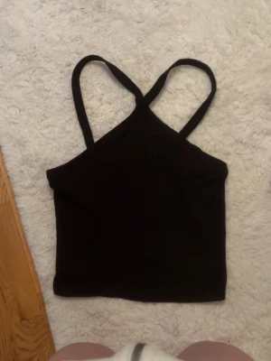 Svart ribbad topp - Säljer en svart ribbad linnetopp med smala axelband som korsas i fram (första bilden är i fram andra bilden är bak). Toppen har en croppad  och är i mjukt stretchigt bomullstyg. 