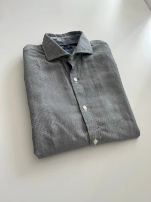 Grå långärmad skjorta från Tommy Hilfiger - Snygg grå skjorta från Tommy Hilfiger med diskret mönster och klassisk krage. Skjortan har vita knappar framtill och på manschetterna, samt lång ärm. Materialet är mjuk bomull som känns skönt mot huden. Perfekt för dig som gillar stilrena och enkla plagg. Oanvänd 