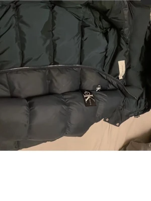 Svart pufferjacka från CK Jeans - Säljer en svart pufferjacka från Calvin Klein Jeans med hög krage och dragkedja. Jackan har en klassisk quiltad design och en tydlig CK-logga på bröstet. Perfekt för kalla dagar och riktigt snygg till streetwear-stilen.