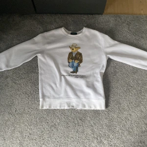 Vit sweatshirt Polo Bear Ralph Lauren - Vit sweatshirt från Polo Ralph Lauren med ikoniskt Polo Bear-tryck på bröstet där björnen har cowboy-outfit. Klassisk rund hals och långa ärmar. Perfekt för dig som gillar streetwear/grisch och vill ha en unik detalj i garderoben.