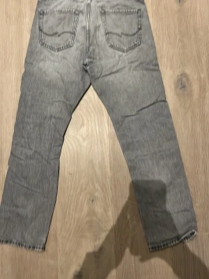 Grå loose fit jeans från Jack & Jones - Säljer ett par grå loose fit jeans från Jack & Jones, modell Loose/Chris. Jeansen har en avslappnad passform, klassiska fem fickor och slitning på ena benet för en cool look. Materialet är bomull och de har en ljusgrå tvättad ton. Har använts mycket 