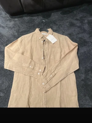 Beige linneskjorta från Gant - Snygg beige skjorta från Gant i luftigt linne. Klassisk button-down krage, bröstficka med diskret Gant-brodyr och vita knappar hela vägen fram. Perfekt till sommarens alla tillfällen och enkel att styla. Självklart äkta ✅, Ställ så mycket frågor ni vill 🌟