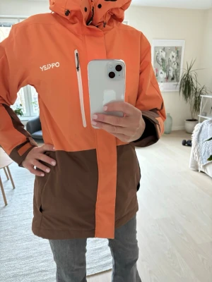 Orange och brun skidjacka från Oakley - Snygg tvåfärgad vindjacka från Oakley i orange och brunt. Jackan har huva med snörning, dragkedja framtill och praktiska fickor. Tillverkad i polyester, perfekt för blåsiga dagar. Stilsäker look med coola detaljer på ärmarna.