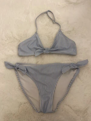 Blåvit randig bikini med knyt - Säljer en söt bikini med blåvita ränder och knytdetaljer både på toppen och trosorna. Bikinin har en halterneck och är gjord i mjukt syntetmaterial som torkar snabbt. Perfekt för strandhäng och poolparty!
