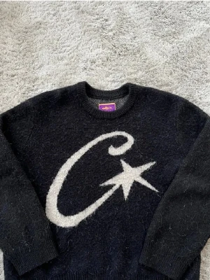 Svart stickad tröja från MS - Svart stickad tröja från MS med stor vit C och stjärna på framsidan. Rund halsringning och långa ärmar. Mjuk och fluffig känsla, perfekt för kyliga dagar. Diskret broderad logga på ena ärmen. Snygg och unik design som sticker ut.