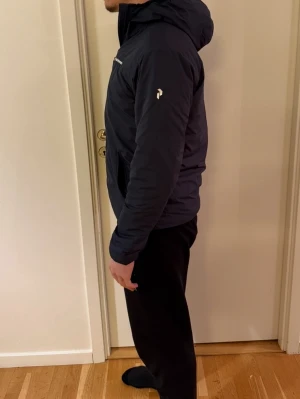 Mörkblå vindjacka Peak Performance - Snygg mörkblå vindjacka från Peak Performance med huva och dragkedja framtill. Jackan har diskreta loggor på bröstet och ärmen, samt två fickor med dragkedja. Perfekt för dig som gillar stilren och sportig look.