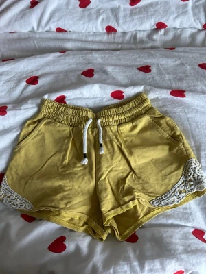 Gula shorts med spets och snörning - Snygga gula shorts från Kappahl med vit snörning i midjan och dekorativ vit spets vid bensluten. Mjukt bomullsmaterial och elastisk midja för skön passform. Perfekta för varma dagar och chill häng.