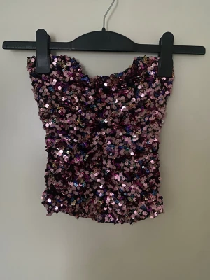 Paljettprydd tubtopp från Zara - Säljer en glittrig tubtopp från Zara i storlek XS, täckt med rosa, lila och blå paljetter som ger ett riktigt partyvibe. Toppen är axelbandslös och har en croppad passform. Perfekt för dig som vill sticka ut med färg och glitter.