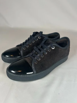 Lanvin captoe - Säljer ett par svarta sneakers från Lanvin med snygg lackad tå och ovandel i mocka. Skorna har snörning med svarta skosnören och rund tå. Sulan är platt och svart, vilket ger en clean look. Perfekta för dig som gillar stilrena och moderna sneakers.
