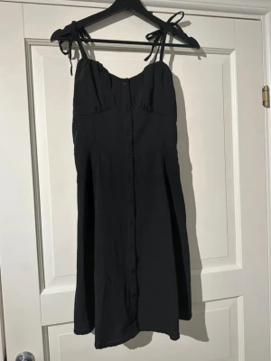 Svart kort klänning med knytband - Svart kort klänning med knytbara axelband och knappar framtill. Klänningen har en smockad rygg och lätt rynkade detaljer vid bysten. Materialet känns som mjuk polyester och modellen är luftig med en normal passform.