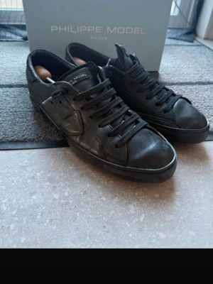 Svarta sneakers från Philippe Model - Säljer ett par svarta Philippe Model sneakers i skinn med klassisk låg profil och rund tå. Skorna har svarta snören, diskret logga på plösen och sidorna samt en platt sula med fiskbensmönster undertill. Perfekta för dig som gillar stilrena och tidlösa sneakers.