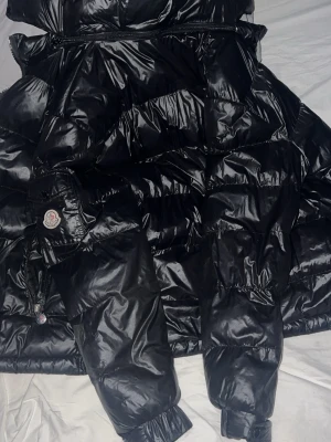 Svart Moncler dunjacka herr L - Säljer en svart glansig dunjacka från Moncler i storlek L. Jackan har klassisk pufferstil, dragkedja framtill, fickor med dragkedja och Moncler-logga på ärmen. Perfekt för kalla dagar och riktigt snygg streetstil.