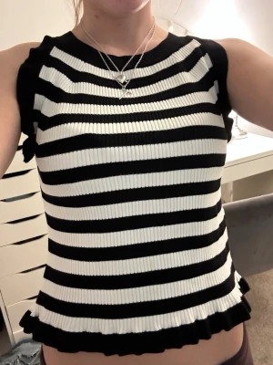 Randig ribbad ärmlös topp - Säljer en svartvit ribbad topp med breda horisontella ränder. Toppen är ärmlös med små volangdetaljer vid axlar och nederkant. Materialet är stretchigt och ger en tight passform. Perfekt att styla med jeans eller kjol för en cool look.