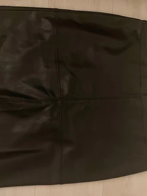 Svart skinnkjol från Soyaconcept - Säljer en svart kort skinnkjol från Soyaconcept i storlek 42. Kjolen har en snygg dragkedja baktill och markerade sömmar som ger en cool look. Perfekt att matcha med boots eller sneakers. 