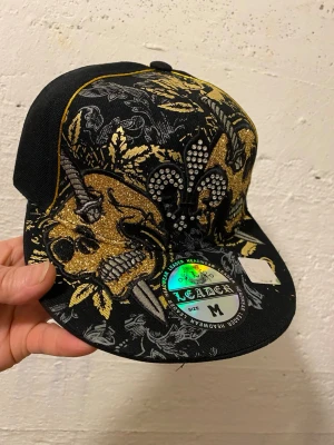 Svart och guld glittrig keps Leader - Säljer en svart keps från Leader med glittriga guldskallar och svärd broderade framtill. Kepsen har även strassdetaljer och ett guldigt liljemärke på baksidan. Cool design med mycket detaljer och kontrasterande färger.