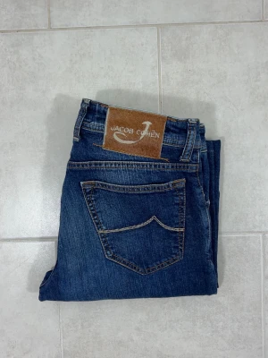 Jacob Cohën Jeans Slim Fit - Jacob Cohën jeans i klassisk mörkblå färg. Mycket bra skick, inga defekter eller anmärkningar. Nypris ca 4000 kr. Material: 98% bomull, 2% elastan. Jeansen är i storlek 31 men små i storleken och passar snarare 29-30. Mått: Midja: 76 cm. Längd (ytterben): 99 cm