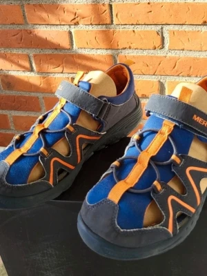 Blå och orange sandaler från Merrell 35 - Säljer ett par sandaler från Merrell i blått och orange med coola detaljer och öppna sidor. Skorna har kardborreband och robust sula, perfekt för äventyr. Materialet är en mix av syntet och mocka med snygga kontrastfärger och grafiska linjer.