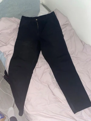 Svarta Carhartt Single Knee Pants - Svarta Carhartt Single Knee Pants med raka ben och klassisk Carhartt-logga på bakfickan. Byxorna har flera praktiska fickor och är gjorda i slitstark bomull med inslag av polyester. Perfekta för dig som gillar streetwear och funktionella detaljer.