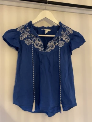 Blå blus med broderade detaljer - Säljer en blå blus med vita broderade blommor vid axlar och halsringning. Blusen har korta ärmar och snörning med tofsar framtill. Materialet känns lätt och luftigt, perfekt för varmare dagar. Snygg och bohemisk stil som sticker ut. Använd 1 gång