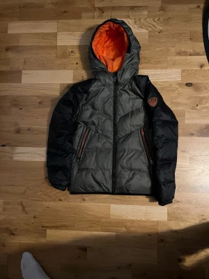 EA7 pufferjacka med huva, svart/grå - Säljer en fet EA7 pufferjacka med svart/grå utsida och orange foder. Jackan har huva, två dragkedjefickor och EA7-logga på ärmen. Perfekt för kalla dagar och riktigt snygg streetstil. Knappt använd, inga defekter, nypris 1699, skriv vid frågor 