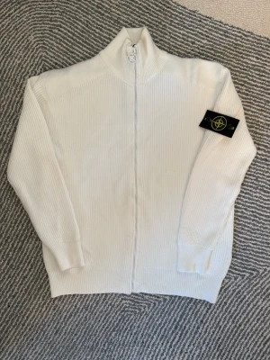 Vit kofta från Stone Island - Säljer en vit ribbstickad kofta från Stone Island med hel dragkedja och ståkrage. Klassisk logotyp-patch på vänster ärm och snyggt designad zip puller. Säljer den för att den är för stor på mig 