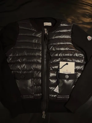  Moncler Cardigan  - Svart pufferjacka från Moncler med quiltad framsida i glansigt material och stickade ärmar. Jackan har två fickor med knappstängning, dragkedja framtill och Moncler-logga på ärmen. Perfekt för dig som vill ha en snygg och modern look.