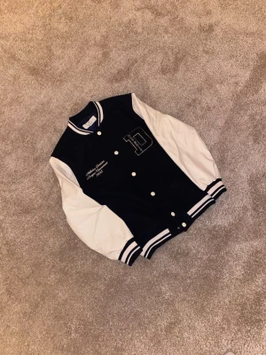 Blåvit bomberjacka från H&M - Snygg blå bomberjacka från H&M med vita ärmar i skinnimitation och broderad text 'Athletics Division League Tournament 1993' samt stort D för Detroit. Jackan har ribbade muddar, tryckknappar och klassisk college-stil. Perfekt för dig som gillar retrovibe!